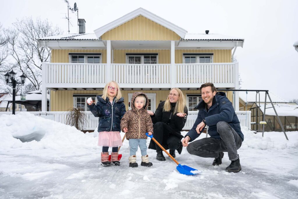 Familjen Linde i Fagersta framför sitt gula och vita trähus. Lite snö på marken och barnen håller i varsin spade för att skotta.